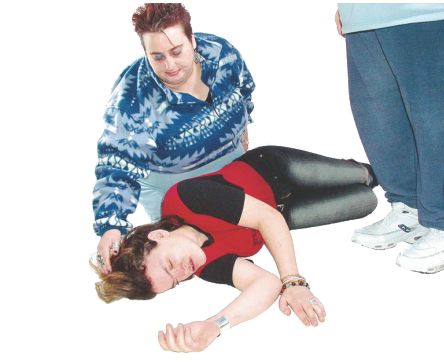 A person is lying on the floor with their eyes closed. Another person is kneeling beside them with their hand on their head. One other person looking over them.