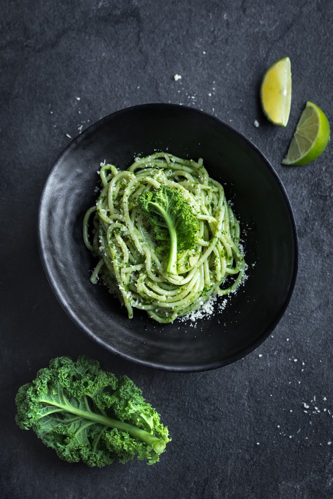 black table with pesto kale pasta in black bowl, lime slices and sprig of kale at the side