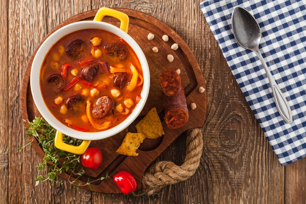Small bowl on wooden board with checked napkin containing cooked chickpeas chorizo and peppers in tomato sauce