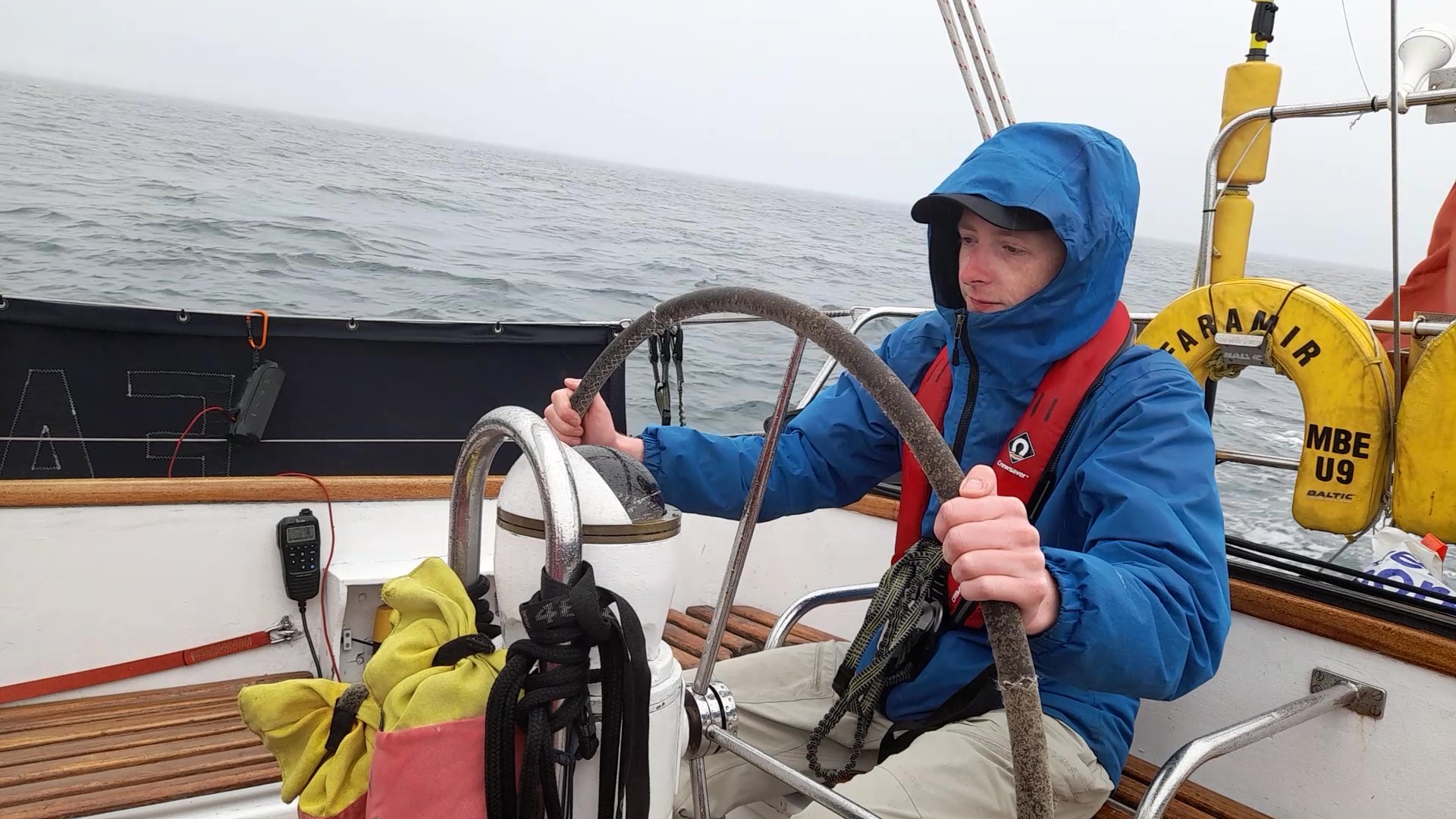 A young man in a blue raincoat at the wheel of a boat, with a grey sea behind