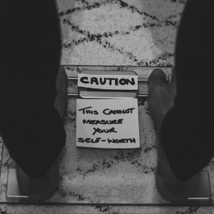 A black and white image of a person standing on weighing scales. On the scales i a piece of paper which says 'caution: this cannot measure your self-worth'