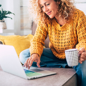 Woman with curly hair in a yellow jumper sits on her sofa with a cup of tea, looking at her laptop