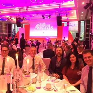 The Health Business awards ceremony stage - people sit at round tables in a pink-lit room, with a backdrop showing the awards' logo