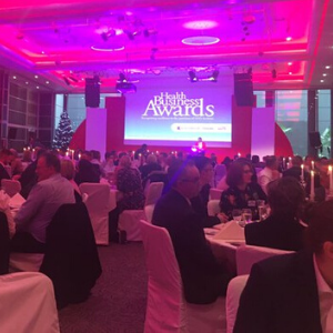 The Health Business awards ceremony stage - people sit at round tables in a pink-lit room, with a backdrop showing the awards' logo