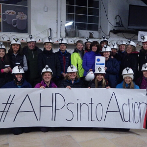 A group of Allied Health Professional staff in hard hats stand hehind a banner that says #AHPsIntoAction