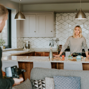 A man stands behind a camera filming a woman in a bright, white decorated kitchen
