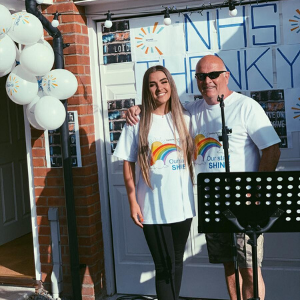 Ellie Mae and her dad wearing SHINE t-shirts in front of their garage door, which is decorated with large letters saying 'NHS Thank You' and balloons. 