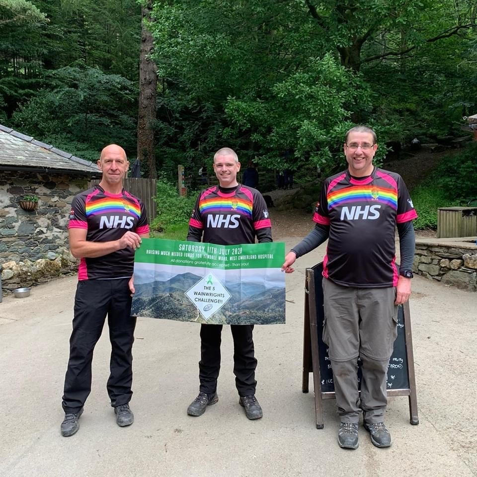 3 men in rugby shirts with rainbows and 'thank you NHS' on stand together in a cap park, holding a banner for the 5 Wainwrights challenge