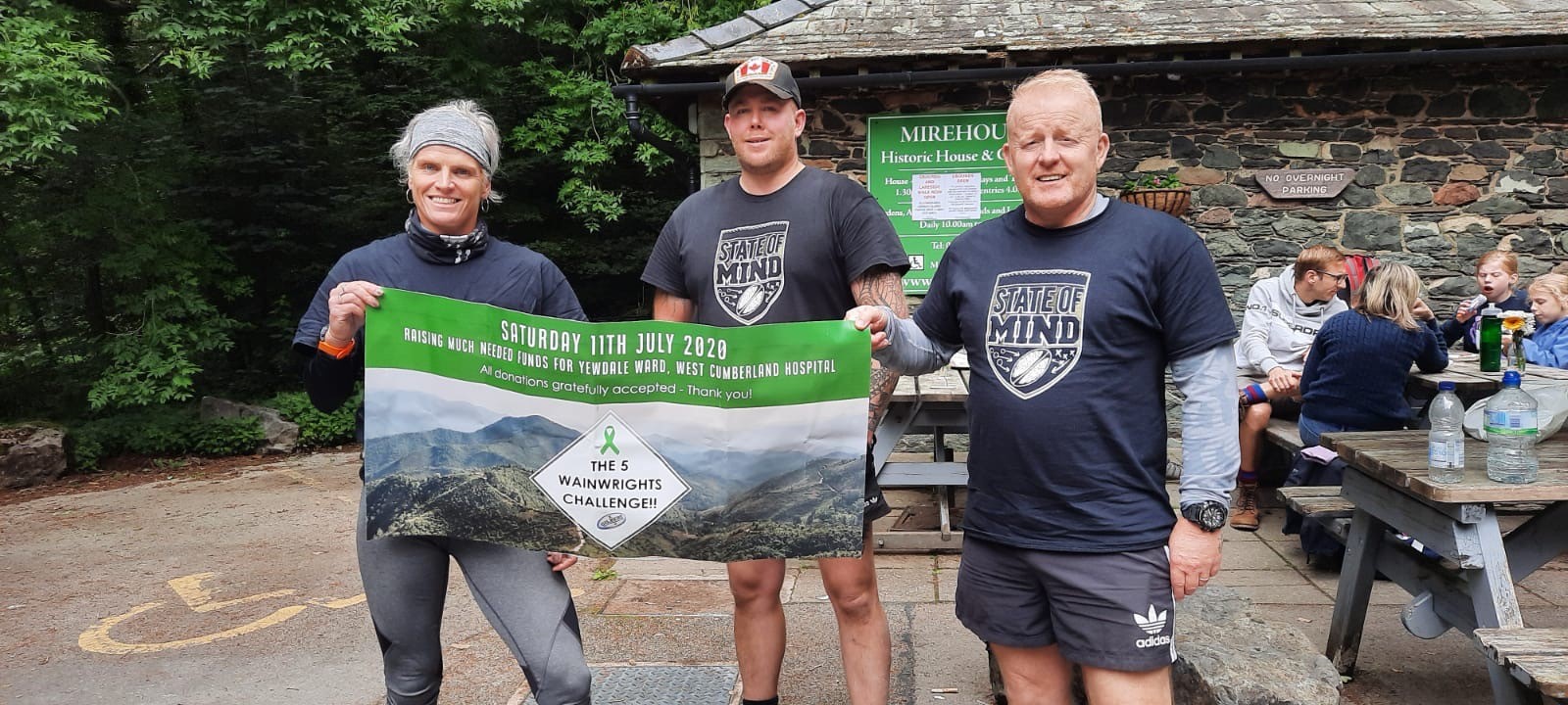 6 men in rugby shirts bearing the State of Mind charity's logo stand together in a cap park, holding a banner for the 5 Wainwrights challenge