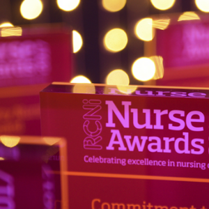 Close-up of a purple perspex award that reads 'RCNi Nurse Awards', against a blurred background of fairy lights and other out-of-focus awards