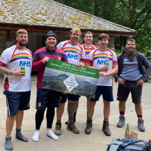 6 men in rugby shirts with rainbows and 'thank you NHS' on stand together in a cap park, holding a banner for the 5 Wainwrights challenge