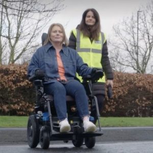 A lady testing a powered wheelchair with a member of the NEDM team following her wearing a high vis jacket.