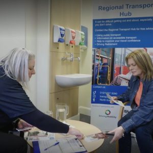 A member of the NEDM team showing a lady an information leaflet. They are seated in the Regional Transport Hub.