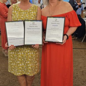 Two members of the team, women with blonde hair in smart dresses, stood smiling and holding their awards