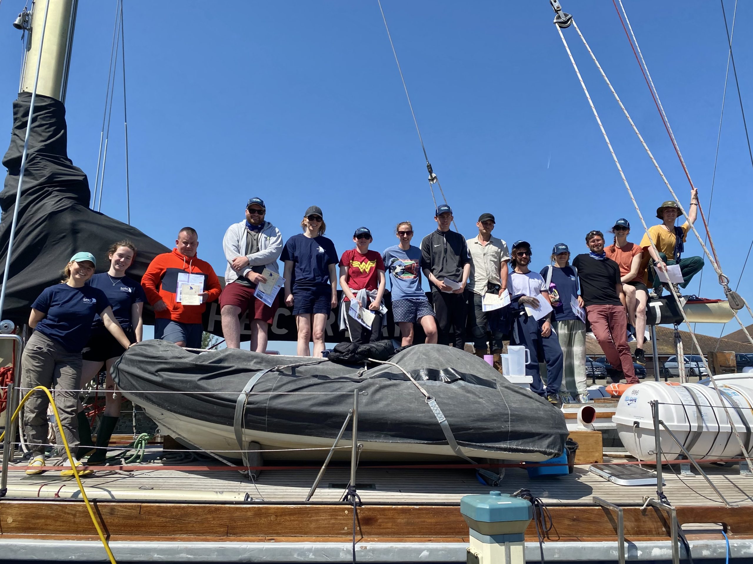 Fourteen men and women standing in a row on a sailing boat holding certificates. 