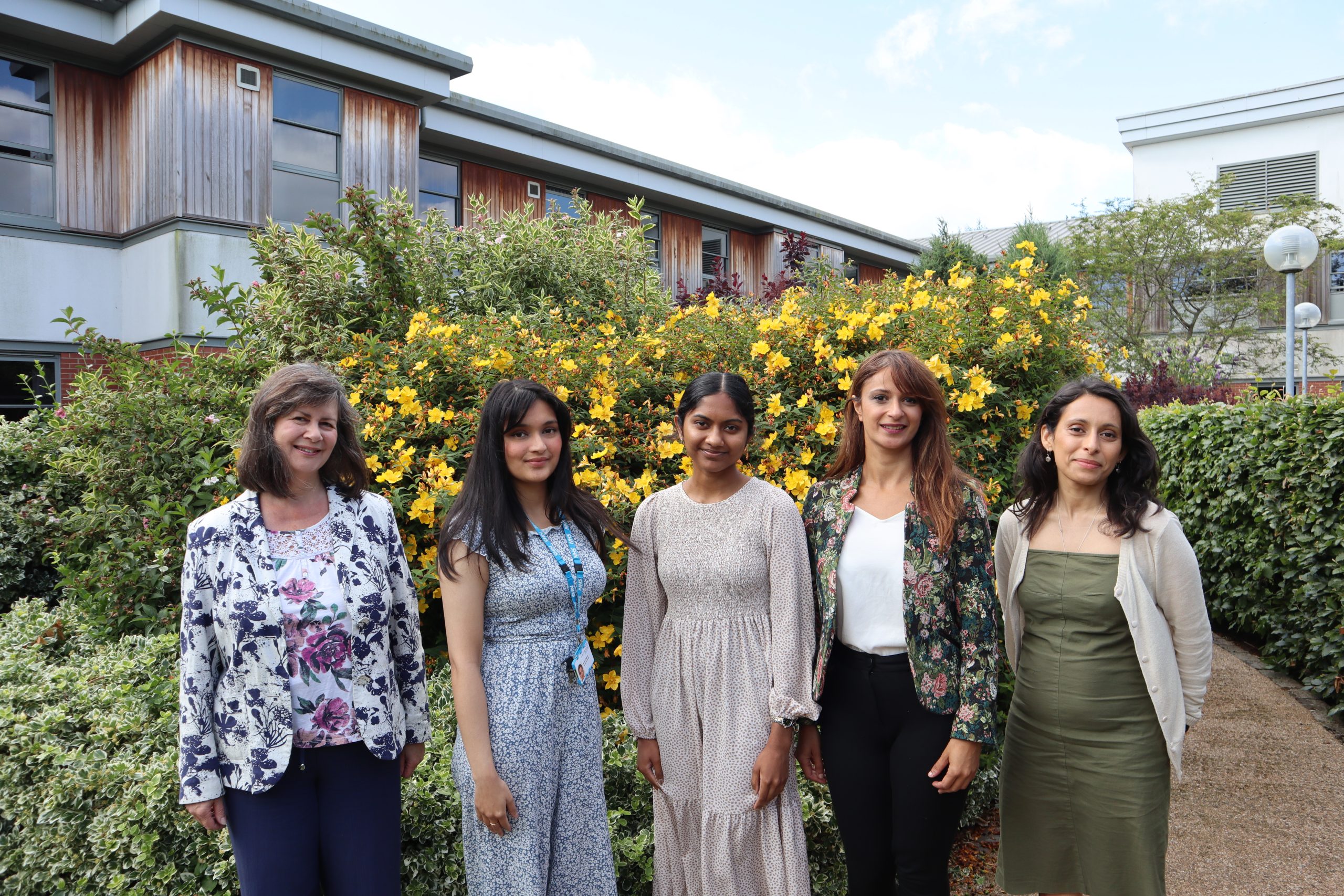 Five smiling women standing outside in front of bush with yellow flowers.