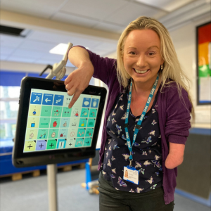 A disabled therapist with a limb difference, wearing black trousers and a purple top with butterflies, smiling, and pointing to a symbol on a Voice Output Communication Aid.