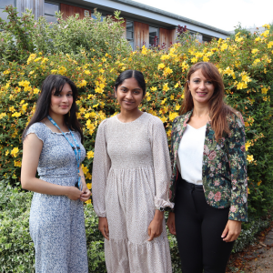 Three smiling women standing outside in front of bush with yellow flowers.