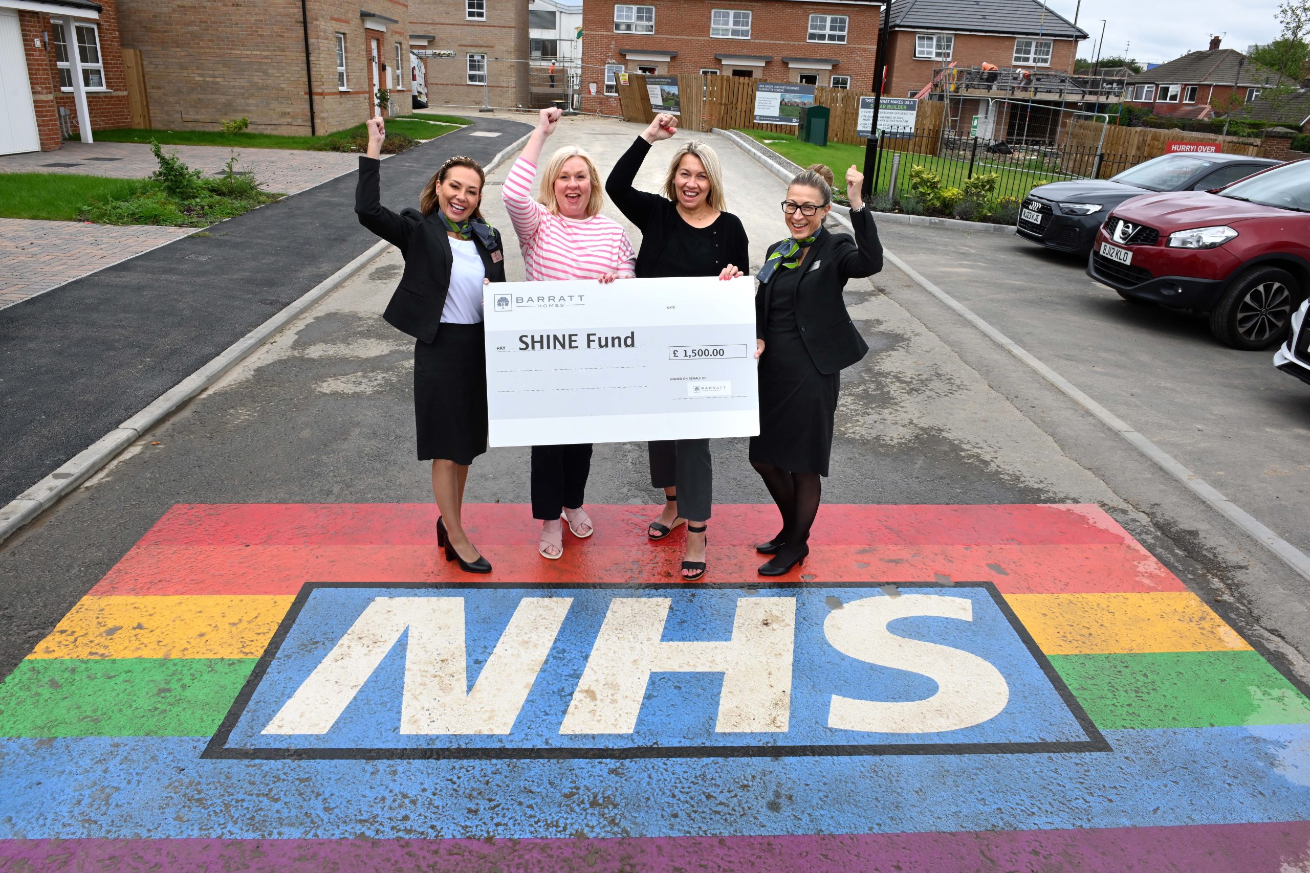 CNTW and Barratt homes staff holding a giant cheque standing on the new painted NHS rainbow at Sycamore Grove