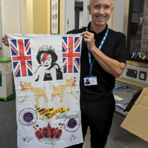Dennis, smiling, hold sup the banner celebratig the Queen's life; it has symbols such as corgis, crowns, and the queen's face