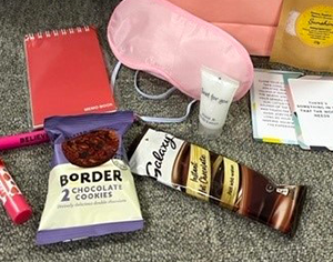 Image of a packet of chocolate cookies, lipbalm, a pen, notebook, sleep mask, hot chocolate packet, hand cream and leaflets