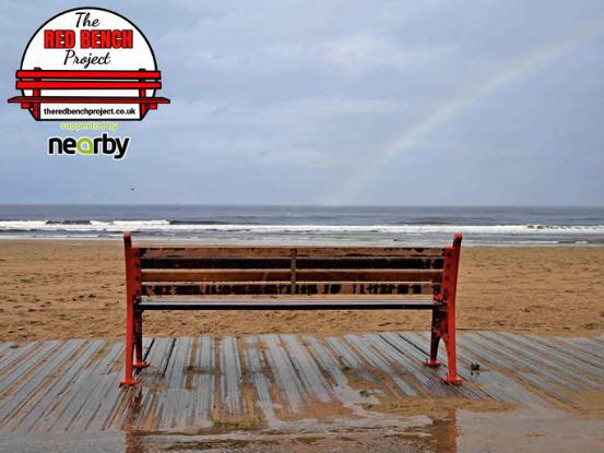 A red bench overlooking the beach with the red bench project logo in the top left-hand side