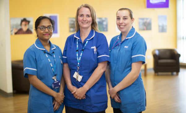 Group photograph of three nurses, wearing blue uniforms, in a ward setting.